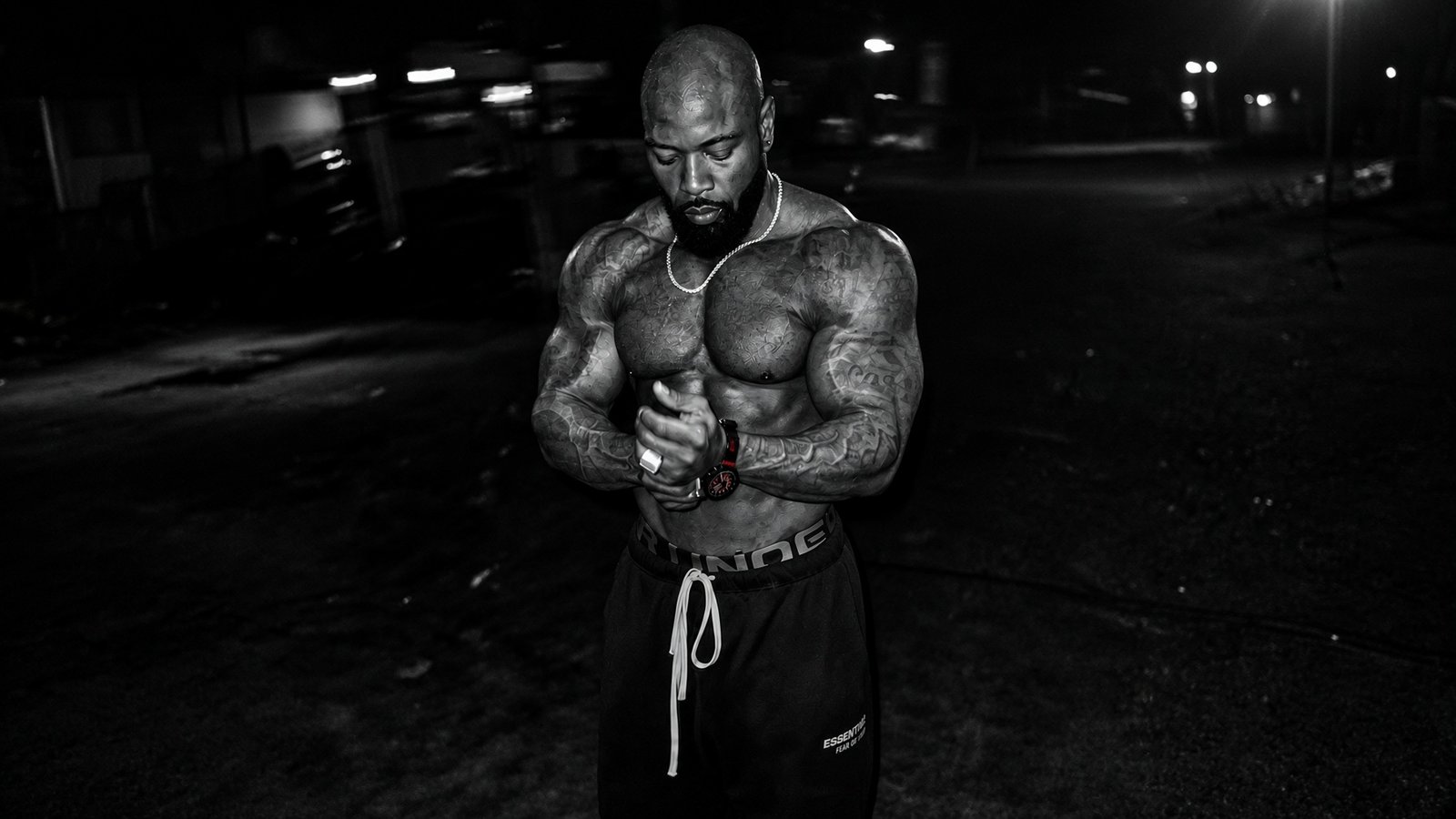 Mike Rashid adjusting his watch in a dark gym, with his tattoos and physique preserved.